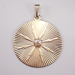 Op de afbeelding: Een gouden hanger met een zonnestralenontwerp en een kleine parel in het midden.