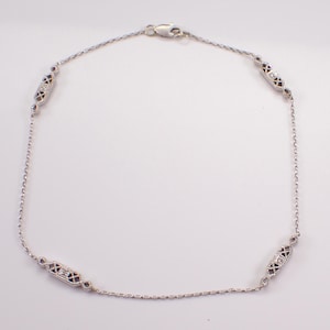 May include: A silver necklace featuring four decorative rectangular sections. Each section has a geometric design with small, clear stones and dark accents. The necklace is on a white background.