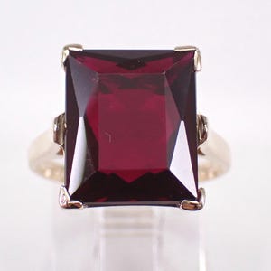 Peut inclure: Une bague en or ornée d'une grande pierre précieuse rouge foncé de forme rectangulaire. La pierre précieuse est au centre, sertie dans un anneau en or avec des détails délicats. Le design de la bague est classique et élégant.