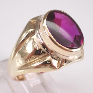 May include: A gold ring featuring a large, oval-cut, deep magenta-colored gemstone set in a smooth, polished gold band. The metal has a high-gloss finish that reflects light, highlighting the rounded shape of the setting and the faceted stone.