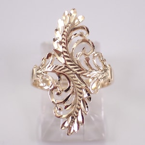 May include: A gold-colored ring with an ornate, openwork design. The ring features a central, vertical leaf-like pattern with intricate details and swirling elements. The ring is displayed on a clear stand.
