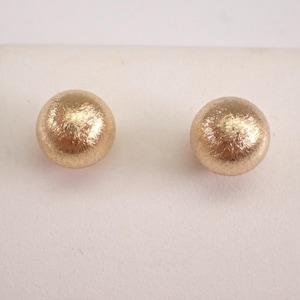 May include: A pair of round stud earrings featuring a textured, brushed gold finish. Each earring is spherical in shape with a matte, metallic surface that catches the light, creating a subtle, multidimensional appearance across the curved surface.