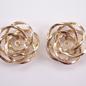 May include: A pair of gold-coloured earrings. Each earring has a circular design made of interwoven loops, forming a floral or rosette pattern. The earrings have a post and backing for secure wear.
