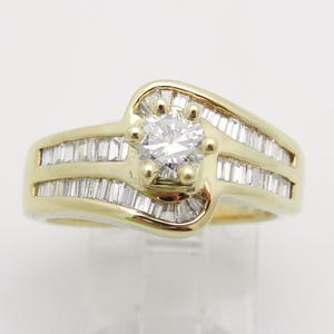 May include: A yellow gold ring featuring a central round brilliant-cut diamond held by four prongs. The band is accented with rows of channel-set baguette-cut diamonds that follow the curved, bypass-style design of the gold metal setting.