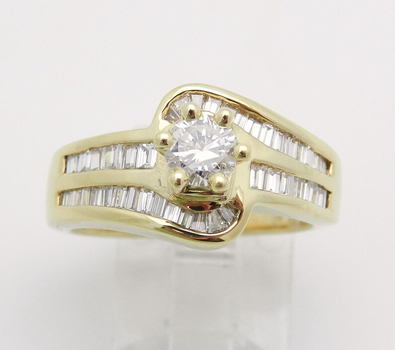 May include: A yellow gold ring featuring a central round brilliant-cut diamond held by four prongs. The band is accented with rows of channel-set baguette-cut diamonds that follow the curved, bypass-style design of the gold metal setting.