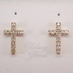 May include: A pair of gold cross-shaped stud earrings, each featuring a vertical row of small, clear, round-cut stones and a horizontal row of similar stones. The text Galaxy Gems is visible in a cursive script between the two earrings.