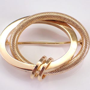 Könnte beinhalten: Goldfarbene Brosche mit ineinandergreifendem Oval-Design. Ein Oval hat eine strukturierte, netzartige Oberfläche, das andere ist glatt. Die Brosche hat eine Nadel auf der Rückseite zum Befestigen. Ein kleiner, goldfarbener Akzent befindet sich unten.