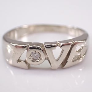 May include: A silver ring with the word "LOVE" cut out. The letter "O" features a small, clear gemstone. The ring is set against a white background, highlighting its polished finish. A simple and elegant piece of jewelry.