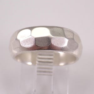 以下が含まれることがあります： テクスチャ加工された幾何学模様のシルバーリング。リングは、ユニークな表面を作り出す一連のファセットを持つ幅広のバンドを備えています。リングは透明なスタンドに展示されており、その洗練された仕上がりを披露しています。