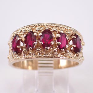 May include: A gold band ring with five oval-shaped red gemstones set in a decorative design.