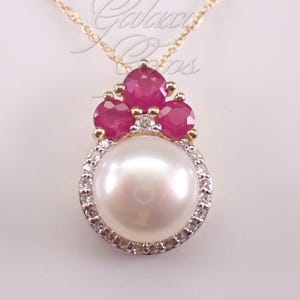 May include: A gold pendant necklace featuring a large, round, white pearl surrounded by a halo of small, clear, sparkling stones. Above the pearl, three round, vibrant pink rubies are set in gold, accented by a single small, clear stone.