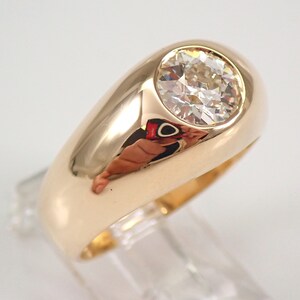 May include: A gold signet ring with a large, round, clear diamond set in the center. The ring has a thick band and a smooth, polished finish.