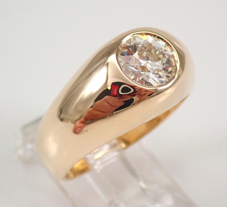 May include: A gold signet ring with a large, round, clear diamond set in the center. The ring has a thick band and a smooth, polished finish.