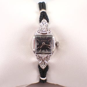 May include: A vintage Wittnauer wristwatch with a black face and silver-tone case. The watch features a decorative silver-tone bezel with diamond accents and a black braided cord band. The watch face displays the numbers 12, 3, 6, and 9.