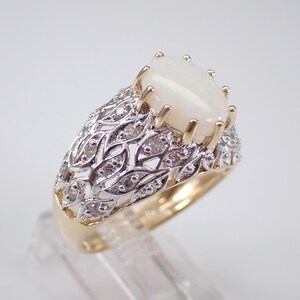 May include: A gold ring featuring a large, oval, white opal gemstone. The ring has a detailed, openwork design with small diamonds set in a silver-coloured metal. The band is gold-coloured.