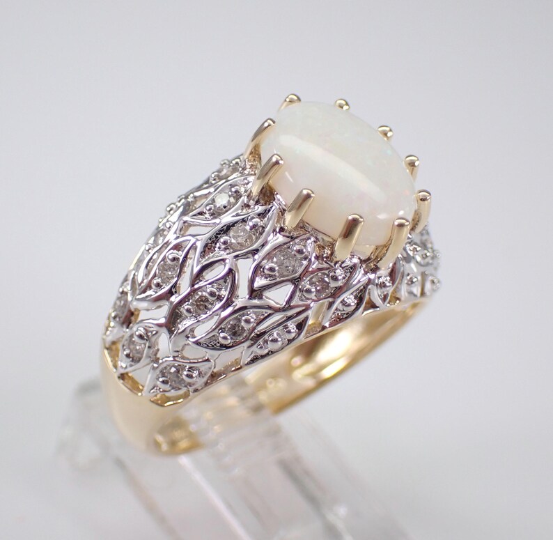 May include: A gold ring featuring a large, oval, white opal gemstone. The ring has a detailed, openwork design with small diamonds set in a silver-coloured metal. The band is gold-coloured.