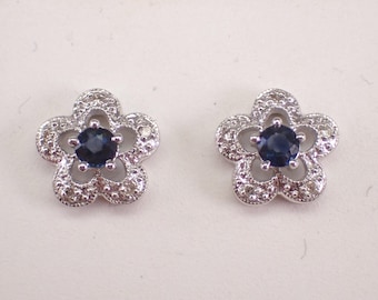 Puede incluir: Un par de pendientes de botón con forma de flor de cinco pétalos. Cada pendiente presenta una piedra central redonda de color azul oscuro rodeada por un marco metálico texturizado, adornado con pequeñas piedras claras y brillantes.