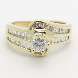 May include: A yellow gold ring featuring a central round brilliant-cut diamond held by four prongs, surrounded by a bypass-style band channel-set with multiple baguette-cut diamonds that extend along the sides of the ring shank.