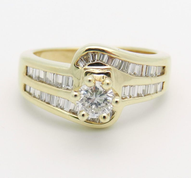 May include: A yellow gold ring featuring a central round brilliant-cut diamond held by four prongs, surrounded by a bypass-style band channel-set with multiple baguette-cut diamonds that extend along the sides of the ring shank.
