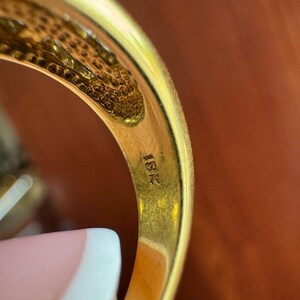 May include: A gold ring with a smooth, polished interior band featuring the stamped text 18K. The metal has a warm, yellow hue, and the inner surface shows a textured, honeycomb-like pattern near the top edge of the band's construction.