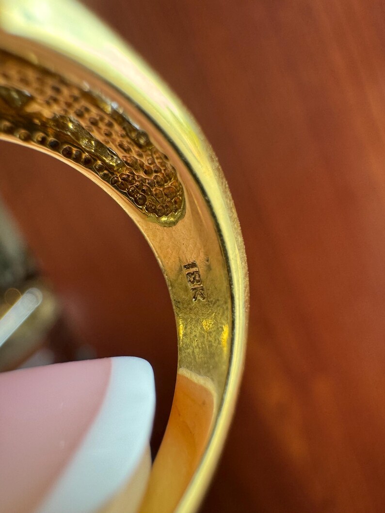 May include: A gold ring with a smooth, polished interior band featuring the stamped text 18K. The metal has a warm, yellow hue, and the inner surface shows a textured, honeycomb-like pattern near the top edge of the band's construction.