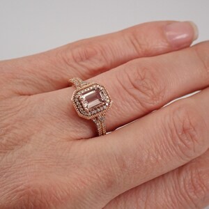 May include: A rose gold ring with a large, rectangular, light pink gemstone surrounded by a halo of smaller diamonds. The ring has a delicate, intricate design.