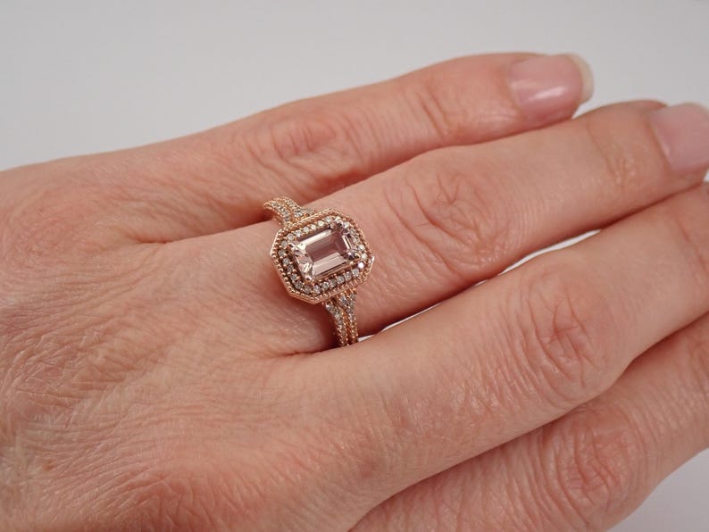 May include: A rose gold ring with a large, rectangular, light pink gemstone surrounded by a halo of smaller diamonds. The ring has a delicate, intricate design.