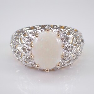 May include: An oval opal gemstone ring. The ring features a large, creamy white opal set in a decorative band with small, clear accent stones. The band has a detailed, openwork design with gold accents.