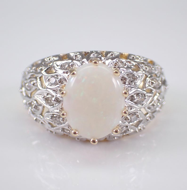May include: An oval opal gemstone ring. The ring features a large, creamy white opal set in a decorative band with small, clear accent stones. The band has a detailed, openwork design with gold accents.