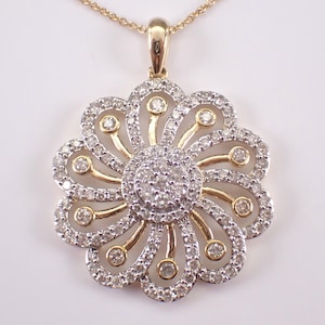 May include: A gold-toned pendant necklace featuring a flower design. The pendant has a central cluster of small diamonds surrounded by a halo of larger diamonds. Gold-toned curved accents connect the center to the outer diamonds, creating a floral pattern.