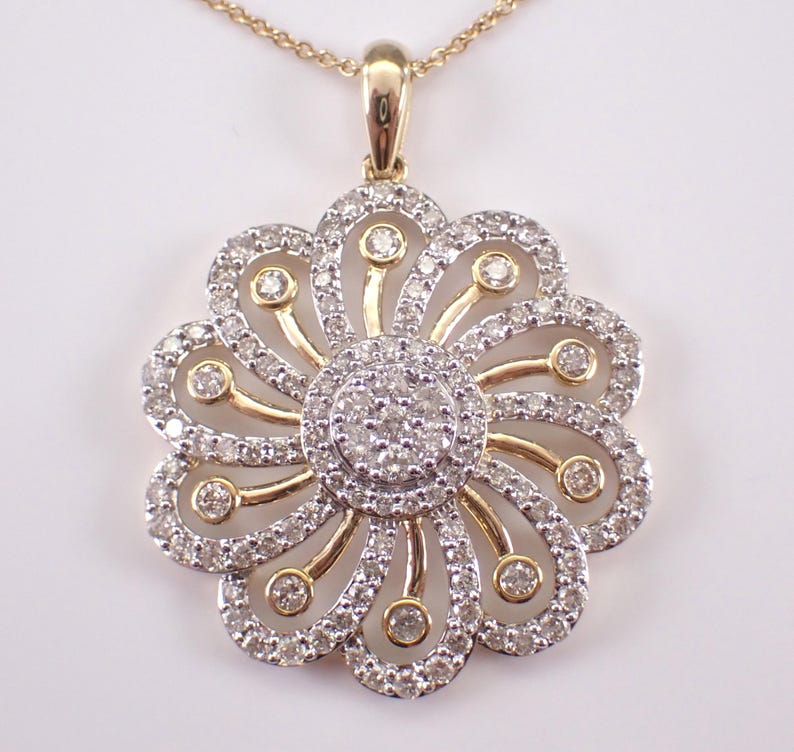 May include: A gold-toned pendant necklace featuring a flower design. The pendant has a central cluster of small diamonds surrounded by a halo of larger diamonds. Gold-toned curved accents connect the center to the outer diamonds, creating a floral pattern.