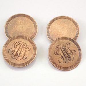 May include: Four round, bronze-colored coasters. Two coasters have a blank surface, while the other two feature an engraved monogram design. The coasters have a raised, concentric ring border. The background is white.