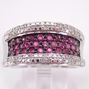 May include: A wide silver ring with a band of small, round, deep pink gemstones set between two rows of smaller, clear gemstones. The ring is displayed on a clear stand against a white background.