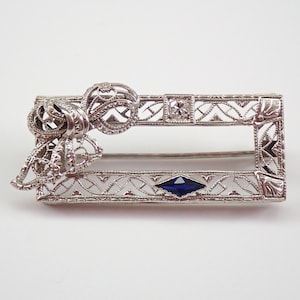 May include: A silver filigree brooch featuring a blue sapphire and a diamond. The brooch is rectangular in shape with a bow on one side.