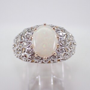 May include: An oval opal ring with a detailed filigree design. The ring features a large, creamy white opal centre stone, surrounded by small diamonds and intricate metalwork. The band has a detailed, openwork pattern.