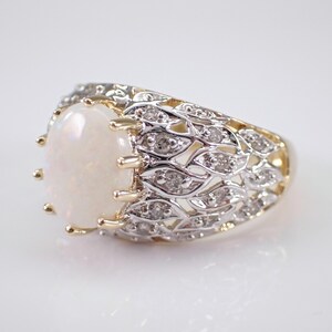 May include: An oval opal ring with a gold band and a decorative silver filigree design. The ring features small diamonds set into the filigree. The opal has a creamy white colour.
