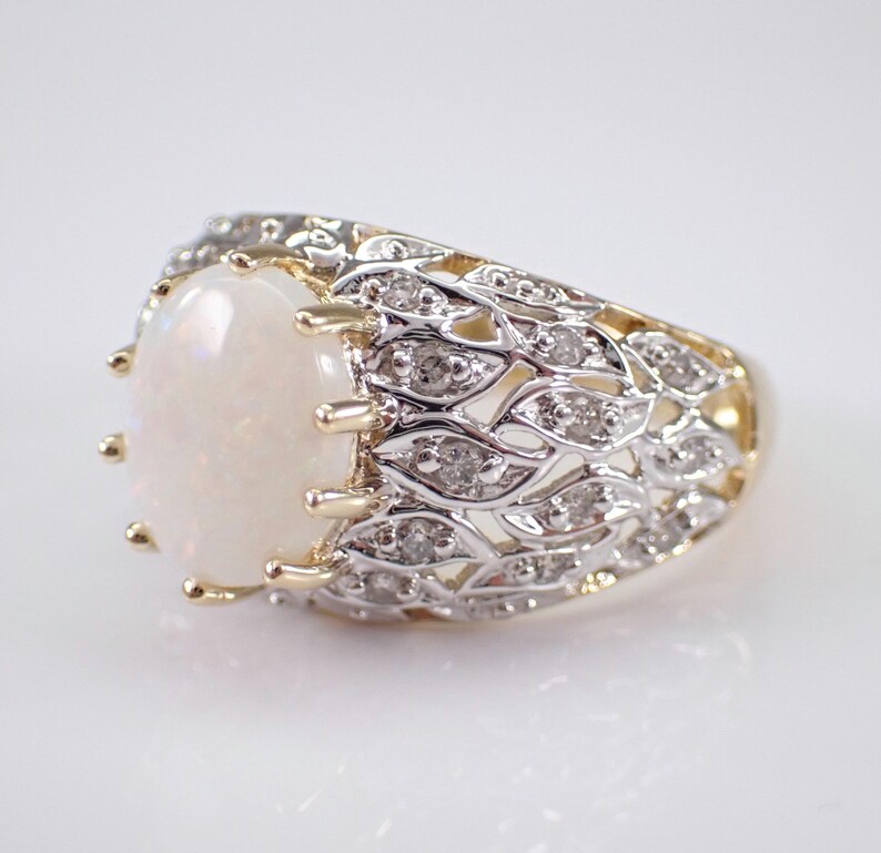 May include: An oval opal ring with a gold band and a decorative silver filigree design. The ring features small diamonds set into the filigree. The opal has a creamy white colour.