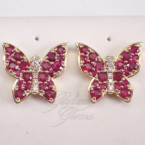 May include: A pair of gold butterfly-shaped earrings featuring wings encrusted with round red rubies and a central body set with small clear white diamonds. The jewellery is displayed on a light-coloured card with the text Galaxy Gems printed below.