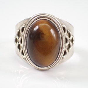 Puede incluir: Un anillo de plata con una piedra preciosa de ojo de tigre marrón engastada en un bisel ovalado. La banda del anillo presenta un diseño decorativo tejido.