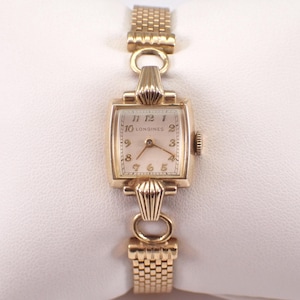 May include: A gold-toned Longines wristwatch with a rectangular face and a linked band. The watch face displays the numbers 1 through 12, with the Longines name printed below the 12. The watch has a small crown on the right side.