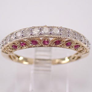 May include: A gold ring with a band featuring a row of clear stones and a row of small red stones. The ring has an ornate, filigree design between the rows of stones. The ring is displayed on a clear stand.