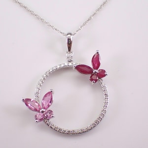 May include: A silver-toned necklace featuring a circular pendant encrusted with small clear stones. Two butterfly-shaped accents are attached to the circle, one set with deep red stones and the other with light pink stones, suspended on a thin chain.
