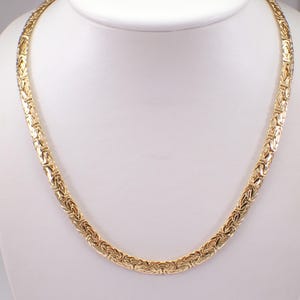 Puede incluir: Un collar dorado con un diseño de cadena bizantina. El collar se exhibe sobre un busto blanco, mostrando sus intrincados eslabones y su estética general. La cadena está hecha de metal dorado.