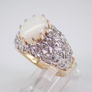 May include: A gold and silver ring with a large, oval, white opal gemstone. The ring has a detailed, openwork design with small diamonds set into the silver band. The opal is held in place by gold prongs.