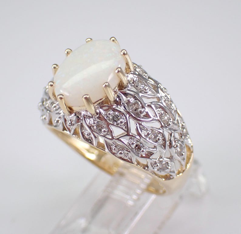 May include: A gold and silver ring with a large, oval, white opal gemstone. The ring has a detailed, openwork design with small diamonds set into the silver band. The opal is held in place by gold prongs.