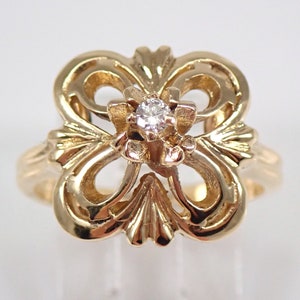 Könnte beinhalten: Ein goldfarbener Ring mit einem verzierten, durchbrochenen Design. Der Ring hat einen kleinen, runden Diamanten in der Mitte. Die Fassung hat ein Blumen- oder stilisiertes Blattmotiv mit polierter Oberfläche. Der Ring ist vor einem schlichten Hintergrund platziert.