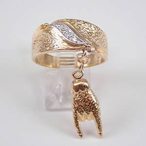 May include: A gold ring with a textured band and a small diamond set in a leaf design. A gold charm in the shape of a hand with the index and pinky fingers extended hangs from the ring.