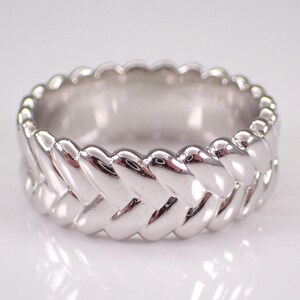 Könnte beinhalten: Ein silberfarbener Ring mit geflochtenem Design. Der Ring hat ein breites Band mit einem sich wiederholenden Muster aus ineinander verwobenen Strängen, wodurch eine strukturierte Oberfläche entsteht. Der Ring ist hochglanzpoliert und reflektiert das Licht.