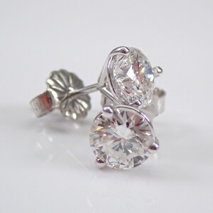 May include: A pair of diamond stud earrings. Each earring features a large, round, brilliant-cut diamond set in a silver or white gold prong setting. The earrings are displayed on a white surface, with the light reflecting off the facets of the diamonds.