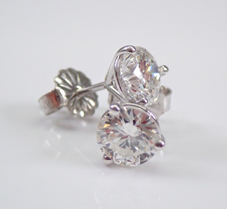 May include: A pair of diamond stud earrings. Each earring features a large, round, brilliant-cut diamond set in a silver or white gold prong setting. The earrings are displayed on a white surface, with the light reflecting off the facets of the diamonds.